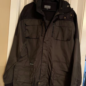 Grå jacka från Euro-Hunt - Säljer en grå jacka från Euro-Hunt Wilderness Wear. Jackan har flera praktiska fickor med lock och dragkedja, samt en huva. Perfekt för utomhusaktiviteter med sin robusta design.