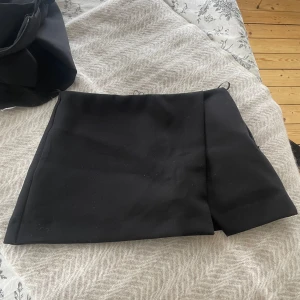 Svart kortkjol från Zara - Snygg svart minikjol från Zara med en stilren design. Kjolen har en liten slits framtill som ger en modern touch. Perfekt för att matcha med olika toppar och accessoarer. Har inbyggda shorts som skorts men kjol baktill också! Helt oanvända och slutat säljas, nu finns det endast sådana med shorts baktill