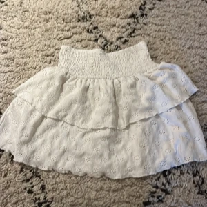 Vit kjol med volang - Charmig vit kjol med volangdetaljer och elastisk midja. Perfekt för en somrig look med sitt lätta och luftiga material. Den har ett subtilt blommönster som ger en romantisk touch.