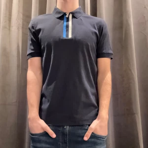 Mörkblå pikétröja från Boss - Snygg mörkblå pikétröja från Boss med en stilren design. Tröjan har korta ärmar och en klassisk krage med en dragkedja i kontrasterande färger. Perfekt för en avslappnad men ändå elegant look.