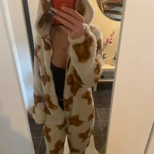 Vit och brun mönstrad onepiece - Mysig vit onepiece med brunt mönster (björnar), perfekt för kalla dagar. Den har en dragkedja framtill som är trasig men går att laga om man är kunnig. och en stor huva för extra komfort med små öron. Materialet ser fluffigt och bekvämt ut, vilket gör den idealisk för avslappnade stunder hemma.