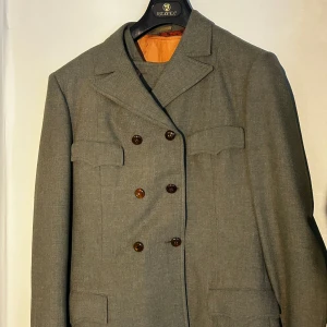 Mörkgrön 3-piece suit - Elegant mörkgrön dubbelknäppt kostym, väst och matchande byxor. Kavajen har en klassisk krage och dekorativa knappar. Västen är ärmlös med samma knäppning. Perfekt för formella tillfällen.