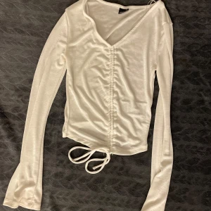 Vit långärmad topp med knyt - Säljer en stilren vit långärmad topp med v-ringning och knytning framtill. Toppen har en åtsittande passform och är tillverkad i ett glansigt material som ger en elegant look.