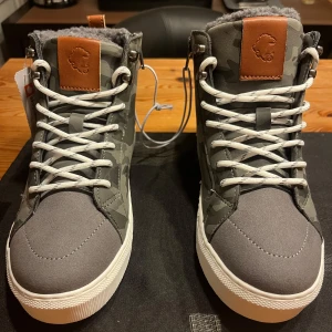 Grå camouflagemönstrade sneakers med snörning - Snygga grå sneakers med camouflagemönster och vita sulor. Skorna har snörning och dragkedja på sidan för enkel på- och avtagning. De har en brun läderdetalj på plösen och baktill. Perfekta för en cool och avslappnad stil.