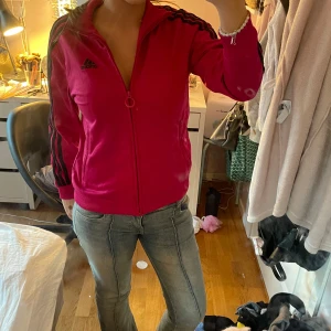 Röd träningsjacka från Adidas - Säljer en snygg röd träningsjacka från Adidas med svarta ränder längs ärmarna. Jackan har dragkedja framtill och en klassisk krage. köpt på second hand men använd väldigt fåtal gånger! lappen är avklippt men skulle gissa på att den är i storlek S eller M