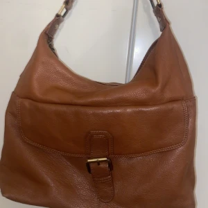 Brun axelväska från M Collection - Snygg brun axelväska i skinn från M Collection. Väskan har en praktisk dragkedja och ett stilrent spänne framtill. Insidan är fodrad med ett ljust tyg med logotyptryck. Perfekt för att bära med sig det nödvändigaste med stil. Äkta skinn. Köpt från Stockholm. Stockholm stil väska. Perfekt för de som gillar att bygga upp sina outfits som gör att de blir ännu snyggare och stiligare💓