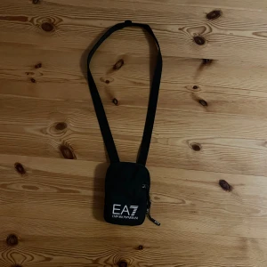 Svart axelväska från Emporio Armani - Snygg svart axelväska från Emporio Armani med justerbar rem. Väskan har en dragkedja och är perfekt för att bära med sig det nödvändigaste. Den är kompakt och stilren med EA7-logga på framsidan. Köpt på NK för 600kr.