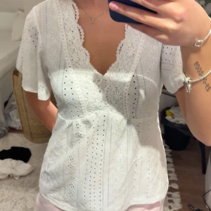 Vit blus med spetsdetaljer - Säljer en söt vit blus med v-ringning och spetsdetaljer. Blusen har korta ärmar och ett fint hålmönster som ger en romantisk känsla. Perfekt för varmare dagar eller sommarens festligheter.