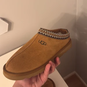 Bruna uggs tasman - Säljer ett par bruna tofflor från UGG med ett snyggt mönster runt öppningen. De är gjorda i mocka och har en mjuk insida för extra komfort. Perfekta för att hålla fötterna varma och sköna. Nästan aldrig använda pga för små 