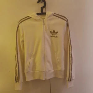 Vit huvtröja från Adidas - Säljer en vit huvtröja från Adidas med klassiska tre ränder längs ärmarna och dragkedja framtill. Tröjan har en huva och två fickor, perfekt för en sportig look. Adidas-loggan pryder bröstet. 🏃‍♂️ Tröjan är i storlek M men är som en S eller XS
