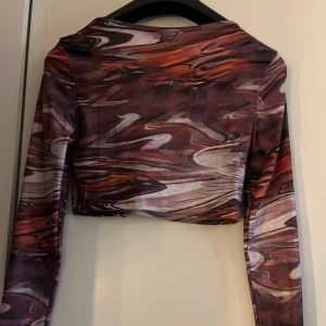 Mönstrad croppad topp - Säljer en cool croppad topp med abstrakt mönster i nyanser av lila, röd och brun. Toppen har långa ärmar och en tight passform, perfekt för en trendig look.