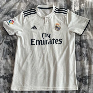 Vit Real Madrid fotbollströja från Adidas - Säljer en vit Real Madrid fotbollströja från Adidas med korta ärmar. Tröjan har klubbens emblem och Fly Emirates-logga på framsidan. Tre svarta ränder pryder axlarna och La Liga-märket sitter på ärmen. Perfekt för alla fotbollsfans! ⚽