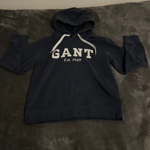Mörkblå hoodie från GANT - Säljer en snygg och bekväm mörkblå hoodie från GANT. Den har en klassisk design med vit text på framsidan och en justerbar huva med vita snören. Perfekt för en avslappnad stil!