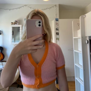 Rosa och orange stickad kofta - Säljer en superfin rosa stickad kofta med orange detaljer. Den är croppad och har korta ärmar, perfekt för vår och sommar. Koftan har knappar framtill och är i ett mjukt och bekvämt material. I nyskick!