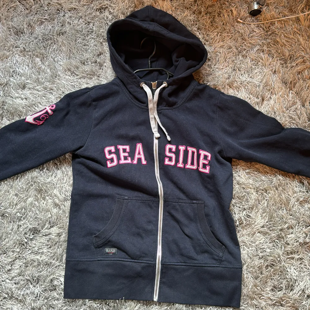 Säljer en navy blå hoodie med dragkedja och texten 'SEASIDE' i rosa vitt på framsidan. Den har en cool ankardetalj på ärmen och en praktisk ficka framtill. Den är väl använd men är fortfarande i ett bra skick 💗🎀. Tröjor & Koftor.