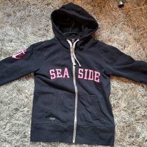 Säljer en navy blå hoodie med dragkedja och texten 'SEASIDE' i rosa vitt på framsidan. Den har en cool ankardetalj på ärmen och en praktisk ficka framtill. Den är väl använd men är fortfarande i ett bra skick 💗🎀