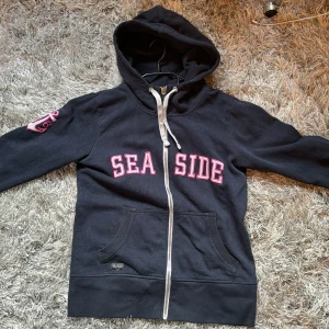 Svart hoodie med texten 'SEASIDE' - Säljer en navy blå hoodie med dragkedja och texten 'SEASIDE' i rosa vitt på framsidan. Den har en cool ankardetalj på ärmen och en praktisk ficka framtill. Den är väl använd men är fortfarande i ett bra skick 💗🎀