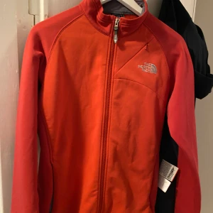 Röd jacka från The North Face - Säljer en snygg röd jacka från The North Face, perfekt för utomhusaktiviteter. Jackan har en dragkedja framtill och en liten bröstficka med dragkedja. Den är en del av Flight Series, vilket gör den idealisk för träning och äventyr. Den har långa ärmar och är i ett syntetmaterial som känns både lätt och bekvämt.