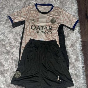 PSG fotbollströja och shorts - Säljer en PSG fotbollströja och shorts i nyskick. Tröjan är kortärmad med ett unikt mönster i beige och svart, och har Qatar Airways som sponsorlogga. Det finns även Jordan-loggan på både tröjan och shortsen. Perfekt för fotbollsfans som vill ha något stilrent och bekvämt!