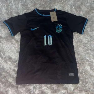 Svart fotbollströja med nummer 10 - Säljer en snygg svart fotbollströja från Nike med korta ärmar. Tröjan har ett coolt blått märke på bröstet och nummer 10 både fram och bak. Den har också texten 'generalen' på ryggen. Perfekt för fotbollsfans som vill sticka ut på planen!