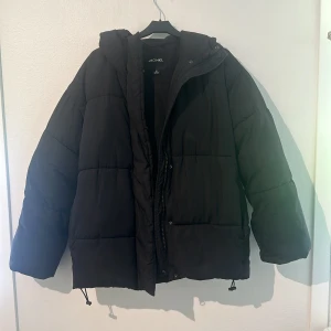 Svart pufferjacka från Monki - Säljer en svart pufferjacka från Monki. Den är perfekt för kalla dagar med sin tjocka vaddering och har en praktisk huva med justerbara snören. Jackan har dragkedja knappar framtill och justerbara snören i nederkanten för att hålla värmen inne. Den är i bra skick endast använd lite förra vintern men nu är den redo för en ny ägare!💓