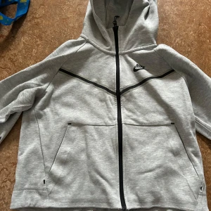Grå hoodie från Nike - Säljer en grå nike kofta  med dragkedja och huva. Den har svarta detaljer och två fickor framtill. Skriv om du är intresserad. 