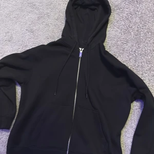 Svart hoodie med dragkedja - Säljer en klassisk svart hoodie med dragkedja och huva. Perfekt för en avslappnad stil. Den har långa ärmar och en praktisk ficka framtill. Passar till alla tillfällen och är ett måste i garderoben.