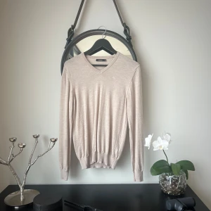 Beige tröja från J.Lindeberg - Säljer en stilren beige tröja från J.Lindeberg. Tröjan har en v-ringad design och långa ärmar, perfekt för en elegant look. Tillverkad i ett mjukt material som ger en bekväm passform. Passar utmärkt till både vardag och mer uppklädda tillfällen. Tröjan kan se lite konstig ut på bild men är helt normal!