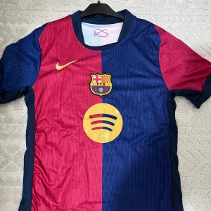 FC Barcelona fotbollströja - Snygg FC Barcelona tröja från Nike i röd och blå med klubbens emblem på bröstet. Tröjan har korta ärmar och ett gult Nike-märke. Perfekt för fans som vill visa sitt stöd.