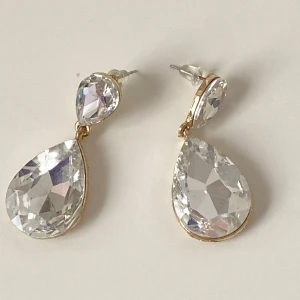 Guldiga örhängen med stora kristaller - Eleganta örhängen i guld med stora, droppformade kristaller. Perfekta för att ge en glittrig touch till din outfit. De har en klassisk design med en klar kristall upptill och en större droppformad kristall nedtill.