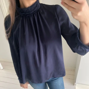 Mörkblå blus med hög krage - Elegant mörkblå blus med hög krage och långa ärmar. Blusen har en lös passform och är tillverkad i ett mjukt, glansigt material som ger en lyxig känsla. Perfekt för både vardag och speciella tillfällen.
