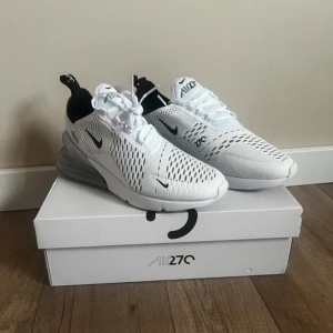 Nike Air Max 270 i vitt - Snygga Nike Air Max 270 sneakers i vitt med svart detalj på hälen. Skorna har en luftig mesh-design och en synlig Air-enhet i hälen för extra komfort. Perfekta för en stilren look.