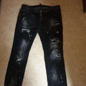 Svarta jeans med slitningar från Dsquared2 - Coola svarta jeans från Dsquared2 med slitningar och färgstänk för en edgy look. De har en straight passform och knappar i gylfen. Perfekta för att ge din outfit en unik touch.  Det står att det e storlek52 men den är inte den är typ 34 elle 42 vet it riktigt  men jag bruka ha på mig M o den passar mig finns 2 håll mellan benen en av de e sytt  (de är dizanade  så)