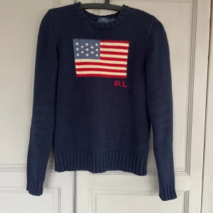 Mörkblå stickad tröja från Ralph Lauren - Säljer en snygg mörkblå stickad tröja från Ralph Lauren med amerikanska flaggan på bröstet. Tröjan har långa ärmar och ribbstickade detaljer vid hals, ärmslut och nederkant. Använd ett fåtal gånger.  Nypris 4895kr