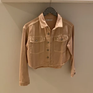 Rosa croppad jacka från Zara - Säljer en beige croppad jacka från Zara med knappar framtill och två bröstfickor. Jackan har långa ärmar och en klassisk krage. Perfekt för en trendig look!