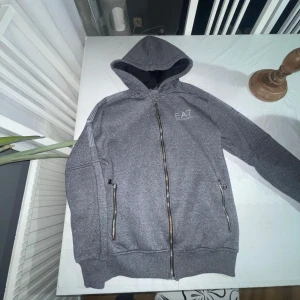 Armani Kofta i Storlek M ( EA7 )  - Säljer en grå huvtröja från EA7 med dragkedja och ribbade muddar. Tröjan har en broderad logga på bröstet och detaljer på ärmen. Perfekt för en casual look. 