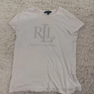 Vit t-shirt från Ralph Lauren - Säljer en klassisk vit t-shirt från Ralph Lauren med RLL-logga på framsidan. T-shirten har korta ärmar och är perfekt för en avslappnad stil. Passar bra till jeans eller shorts.
