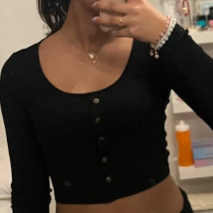 Svart ribbad croppad topp - Snygg svart ribbad topp med knappar framtill. Den är croppad och har långa ärmar, perfekt för en trendig look.