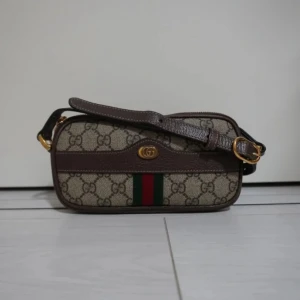 Beige och brun axelväska från Gucci - Snygg axelväska från Gucci i beige och brunt med det klassiska GG-mönstret. Väskan har en justerbar axelrem och detaljer i guld. Den har en dragkedja upptill och de ikoniska gröna och röda ränderna framtill. Perfekt för att ge din outfit en lyxig touch.