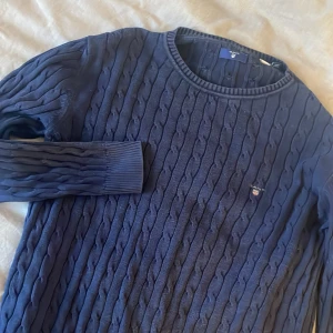 Mörkblå stickad tröja från GANT - Säljer en snygg mörkblå stickad tröja från GANT med kabelstickat mönster. Tröjan har långa ärmar och ribbade muddar vid ärmslut och hals. Perfekt för kyligare dagar!
