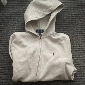 Grå hoodie från Ralph Lauren - Säljer en stilren grå hoodie från Ralph Lauren med dragkedja och den klassiska loggan på bröstet. Mycket bra skick men ganska så kort i ärmarna 