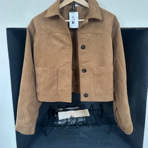 Brun mockajacka med knappar - Snygg brun mockajacka med knappar framtill och två stora fickor. Jackan har en klassisk krage och är croppad i modellen. Perfekt för höst och vår.