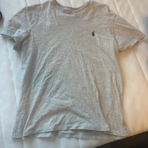 Grå t-shirt från Ralph Lauren - Säljer en klassisk grå t-shirt från Ralph Lauren med det ikoniska logotypen broderad på bröstet. T-shirten är tillverkad i mjuk bomull. Perfekt för en avslappnad stil. Lite utvättad men annars bra.