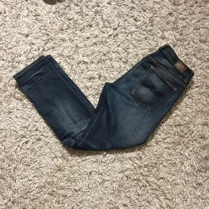 Mörkblå jeans från Nudie modell grim trim  - Snygga mörkblå jeans från Nudie modell grim trim, med klassisk femficksdesign och orange sömmar. De har en slim/straight passform och är perfekta för en avslappnad stil. Jeansen har en knappgylf och är tillverkade i slitstarkt denim. Riktigt fet färg på dem med mörbrodering på bakfickorna. Strlk 32 / 32, mått kan skickas privat.  Gratis frakt just nu så passa på! Postas antigen samma dag som köp eller dagen därpå🙌💯