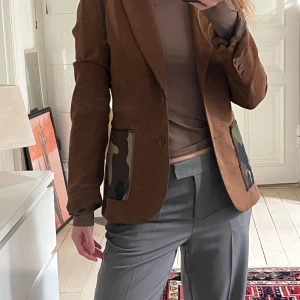 Brun kavaj med camouflage-detaljer - Snygg brun kavaj med unika camouflage-detaljer på fickorna. Kavajen har en klassisk passform med knappar framtill och långa ärmar. Perfekt för att ge en stilren look en twist. 🤎