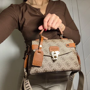 Beige och brun handväska från Guess - Snygg handväska från Guess i beige och brunt med logotypmönster. Väskan har en justerbar axelrem och ett handtag i brunt skinn. Den stängs med ett metallås framtill och har fina detaljer i skinn. Perfekt för att ge din outfit en stilfull touch.