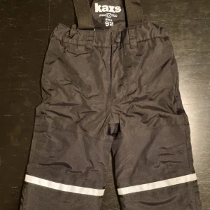 Svarta täckbyxor från Kaxs - Svarta vinterbyxor från Kaxs med justerbara och avtagbara hängslen och reflexdetaljer på benen. Snölås vid benslut. Justerbar midja. Perfekta för vinteraktiviteter med sitt slitstarka material och bekväma passform. 