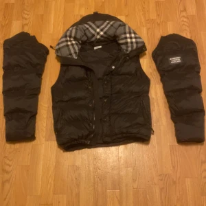 Burberry jacka/väst - Säljer en snygg svart dunjacka från Burberry med avtagbara ärmar. Jackan har en klassisk rutig huva och dragkedja framtill. Perfekt för kyliga dagar när du vill hålla stilen. Storlek L men sitter mer som M är justerbar i storleken också. Kan bytas mot en mindre storlek eller nått annat