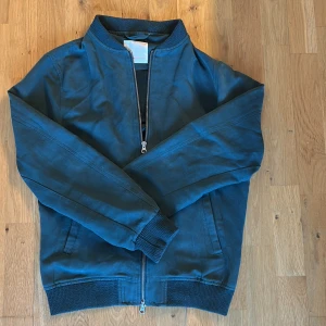 Mörkgrön vår jacka - Snygg mörkgrön jacka i mocka med dragkedja och ribbade muddar. Perfekt för en stilren look. Utmärkt att ha nu på våren då den är tunn och stilren!