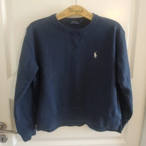 Kortare sweatshirt med slits, Ralph Lauren - Riktigt snygg sweatshirt från Ralph Lauren med slits, något längre bak än fram. Mörkt blåmelerad med vit logga. Mjuk fleecekänsla på insidan. Stl S, kortare modell, drar eventuellt lite mot XS, se mått. Mycket fint skick, inga hål eller fläckar.  Mått: Armhåla till armhåla 51cm, bredd längst ner 47cm, längd från mitt på axeln framsida 50cm och baksida 53cm.